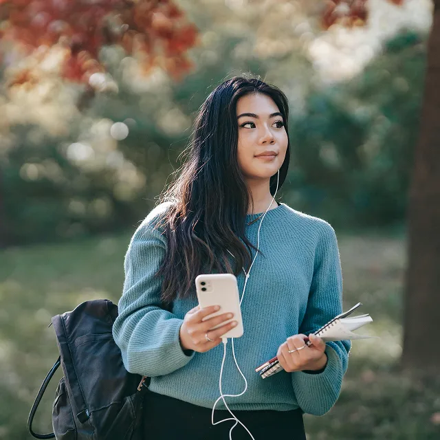 Young woman in a teal sweater holding a smartphone, notebook, and pens, listening to music with earphones, standing outdoors with blurred autumn trees in the background.