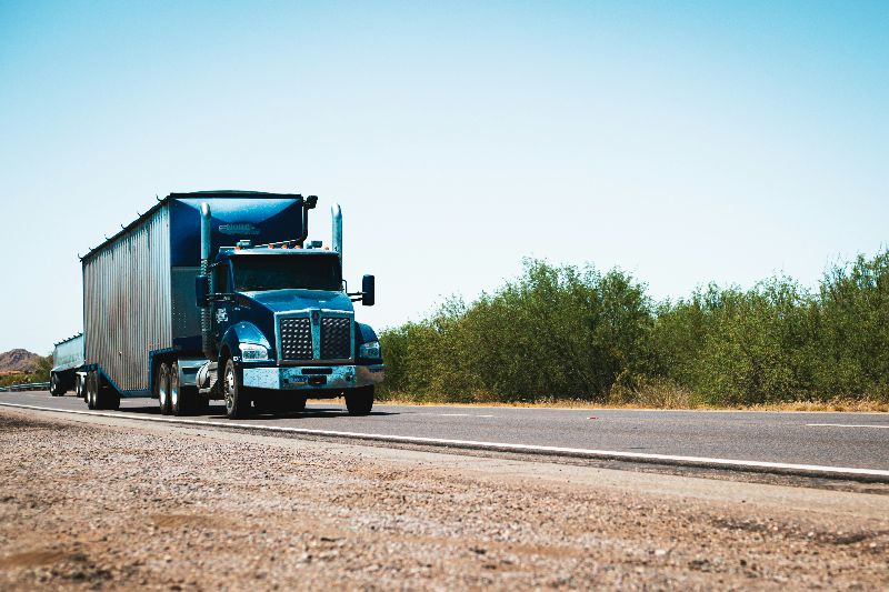 Blue semi truck pulling long trailer driving on rural highway with desert landscape and clear sky background.