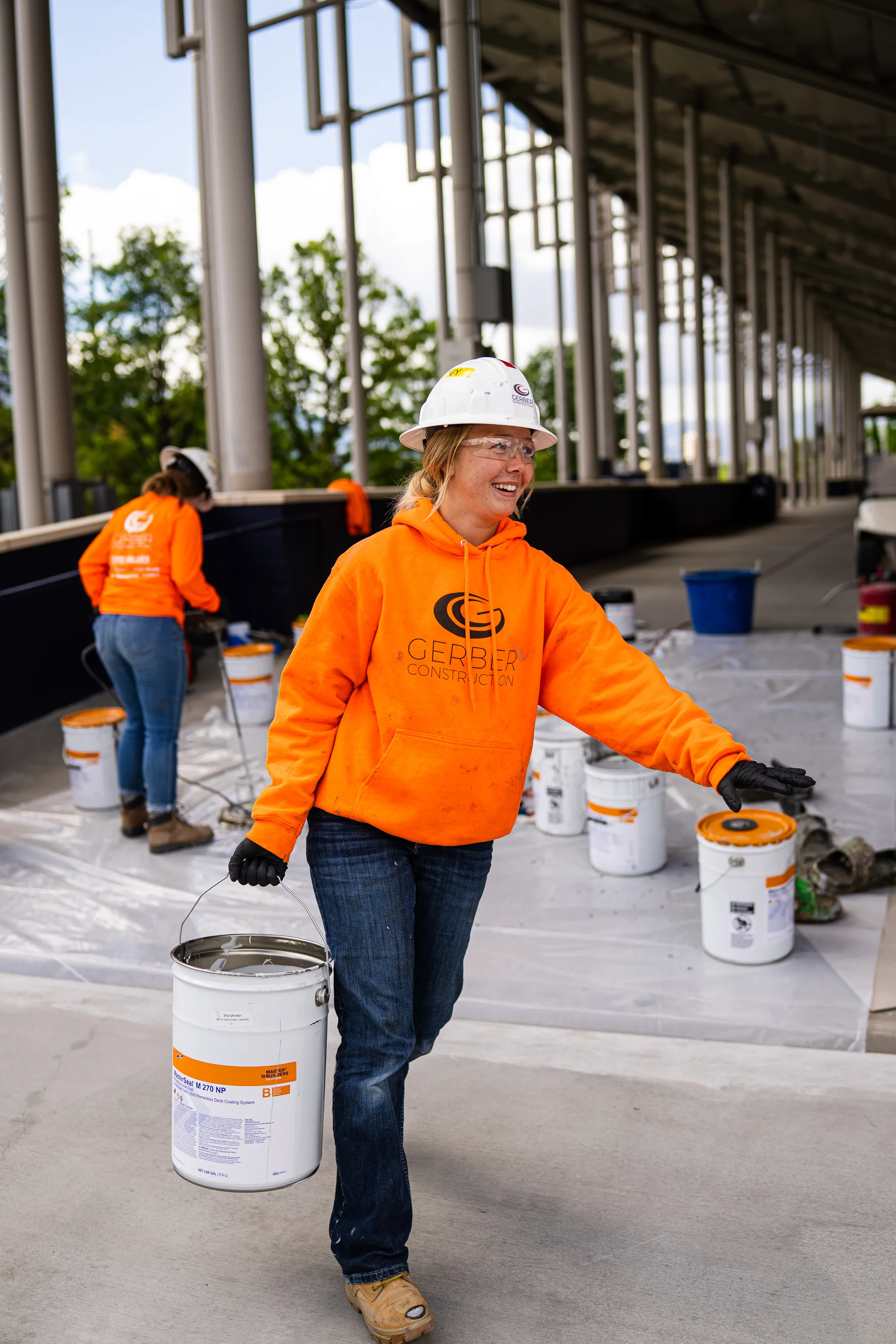 A smiling Gerber crew member carries a bucket of deck coating across a prepared concrete deck.