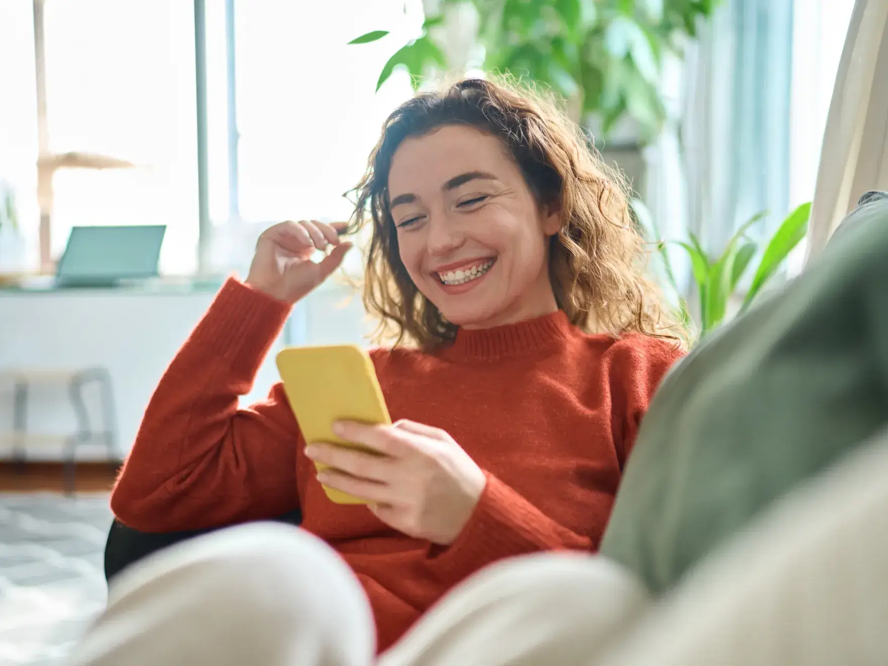 A person in a red sweater smiles while looking at their phone, sitting on a couch.
