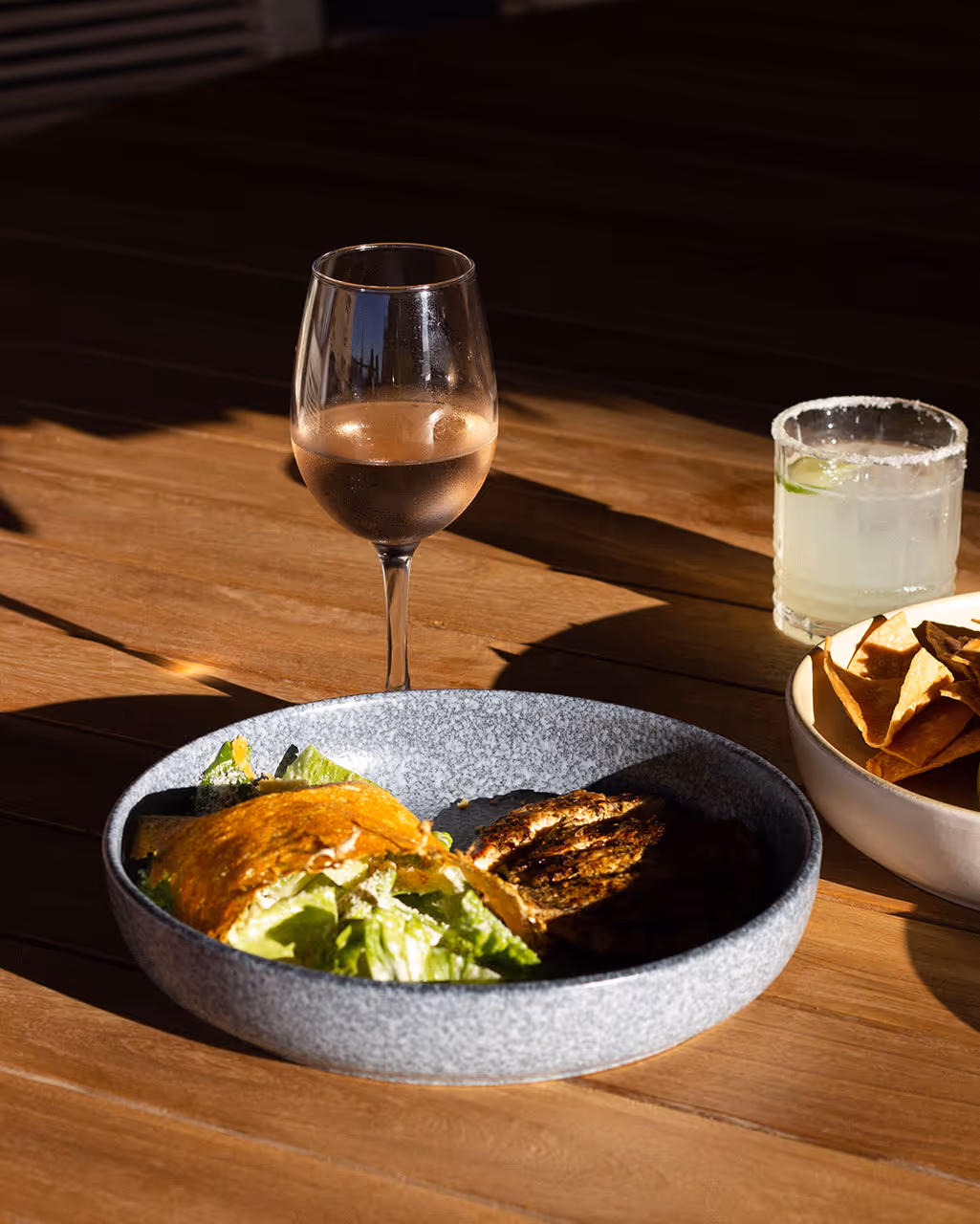 A gray bowl containing a taco with lettuce and a side of grilled chicken, a glass of rosé wine, a cocktail with lime, and a bowl of tortilla chips on a wooden table. The setting suggests a relaxed beach vibe in Baja California, perfect for an adventure.