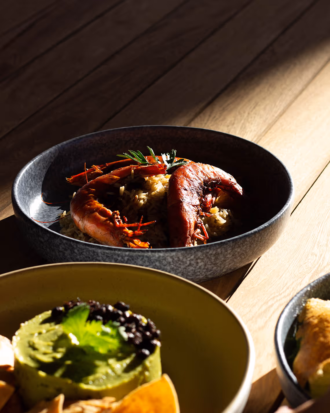 A close-up of a dark bowl containing large shrimp garnished with herbs and spices, placed on a wooden table. Other colorful dishes are partially visible in the foreground, offering a variety of textures and ingredients.