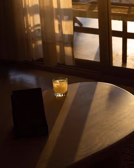 A glass of yellow drink on a wooden table with a black menu beside it. Soft sunlight filters through sheer curtains, creating long shadows on the surface.