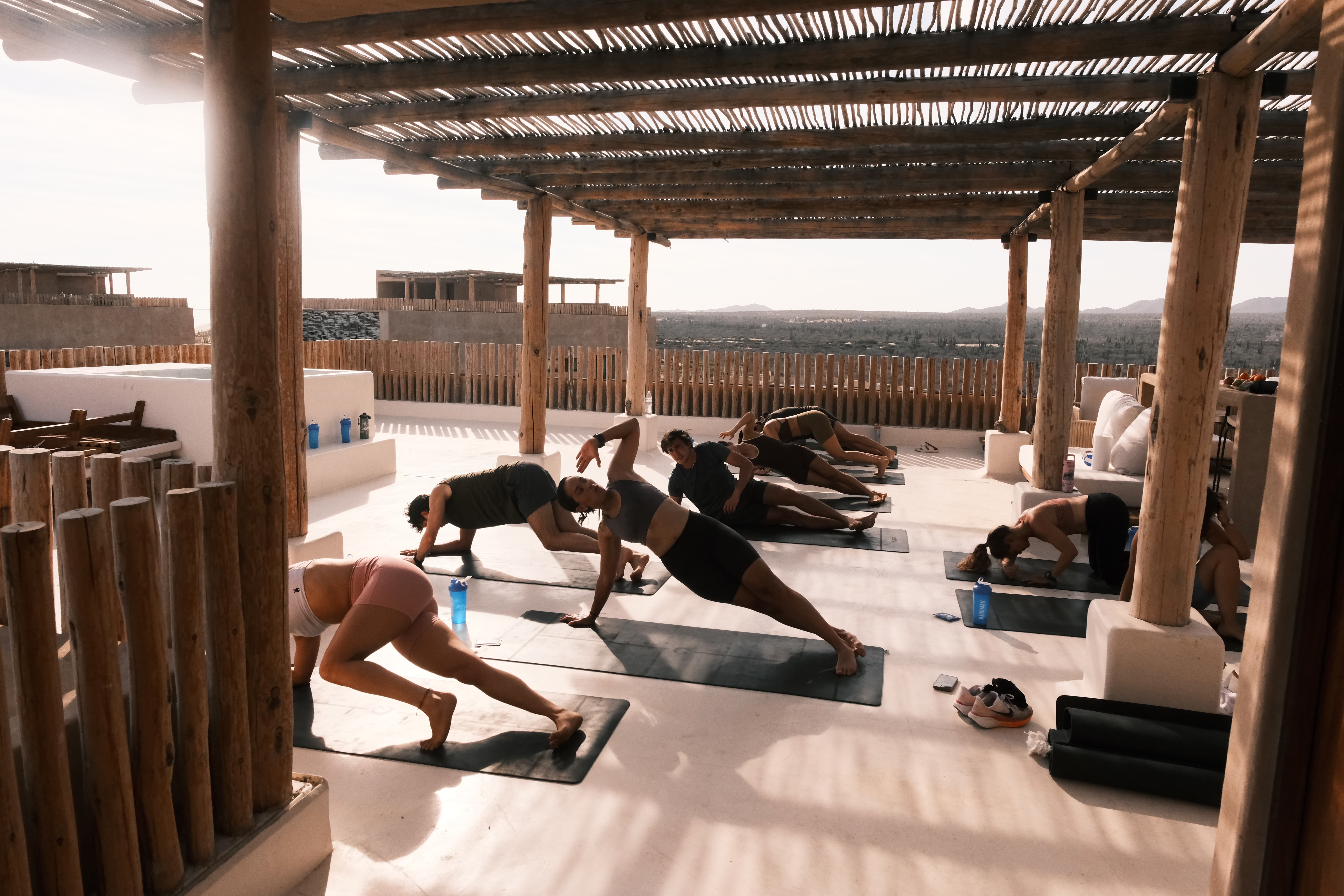 A group of individuals practicing yoga on mats outdoors during sunset near the ocean. Palm trees are visible, creating a serene atmosphere. The scene conveys a sense of adventure and tranquility in Baja California.