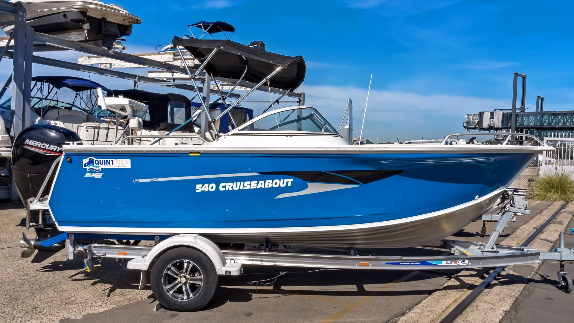A blue "540 Cruiseabout" motorboat on a trailer, parked on a concrete surface. The boat is sleek and modern, with a sunny, clear sky above.