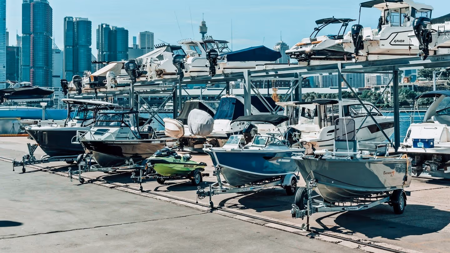 Boats and jet skis on trailers and racks at a marina, with a city skyline in the background. The scene is bright and bustling, conveying a summery, nautical feel.