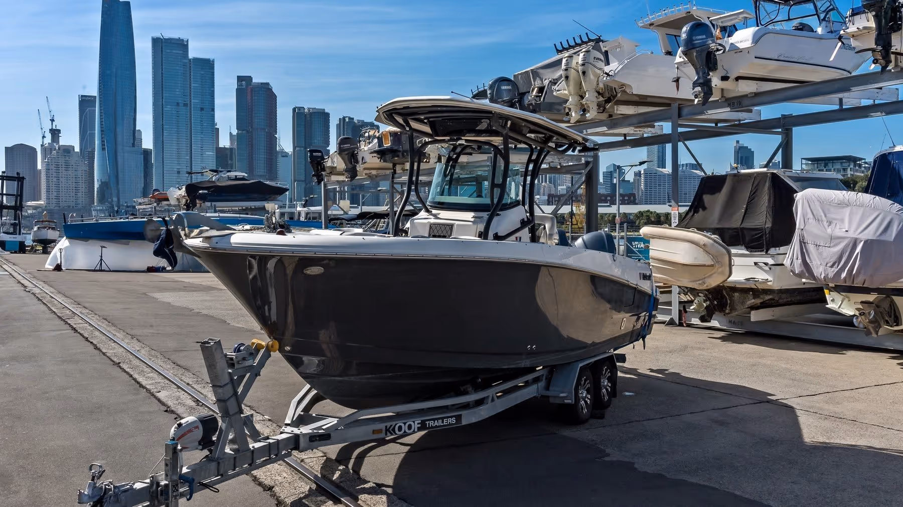 A sleek motorboat on a trailer in an urban marina, with a city skyline backdrop. Other boats are stacked nearby, under a clear blue sky.