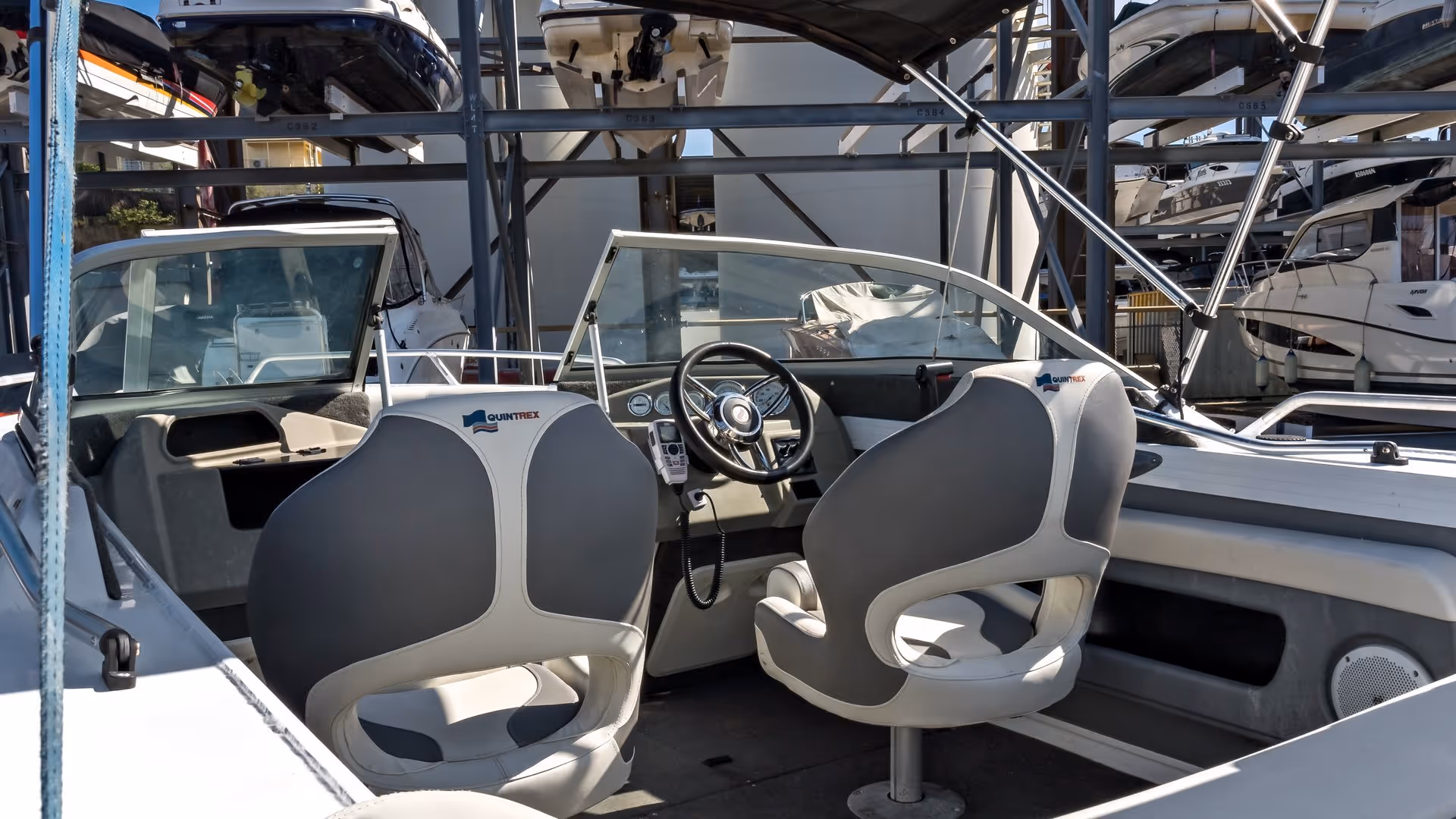 Interior of a small motorboat shows two gray and white seats facing a steering wheel, surrounded by stored boats on stacked racks in a sunny marina.