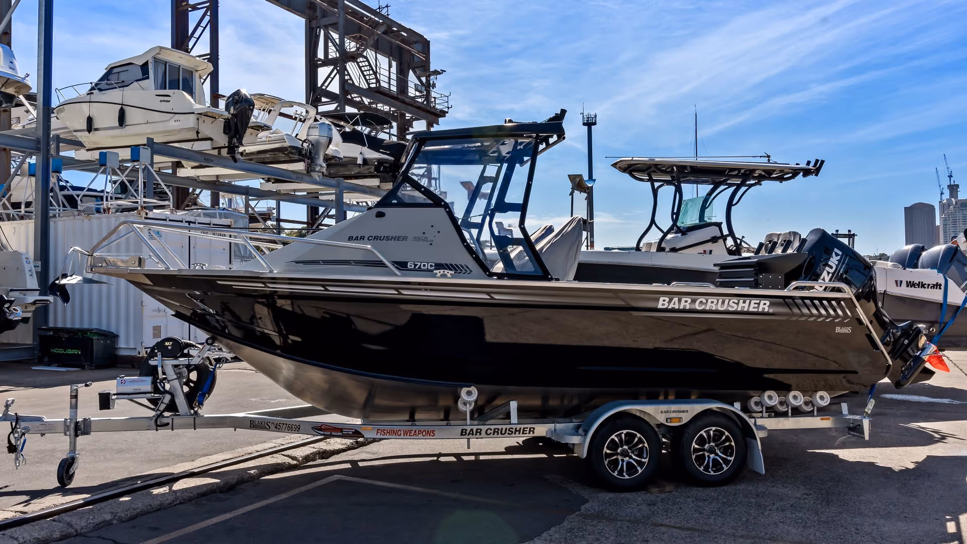 A sleek black Bar Crusher boat on a trailer in a marina, surrounded by stacked boats and a clear blue sky. The scene feels industrial yet serene.