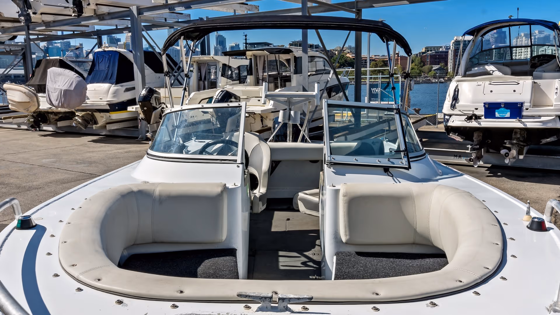Bow view of a white speedboat with cushioned seating, docked among various boats on a sunny day. City skyline and water are visible in the background.