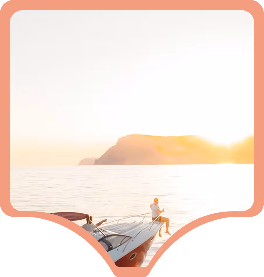 A person relaxes at the bow of a yacht during a serene sunset. The sun sets near a distant island, casting a warm glow over the calm sea.