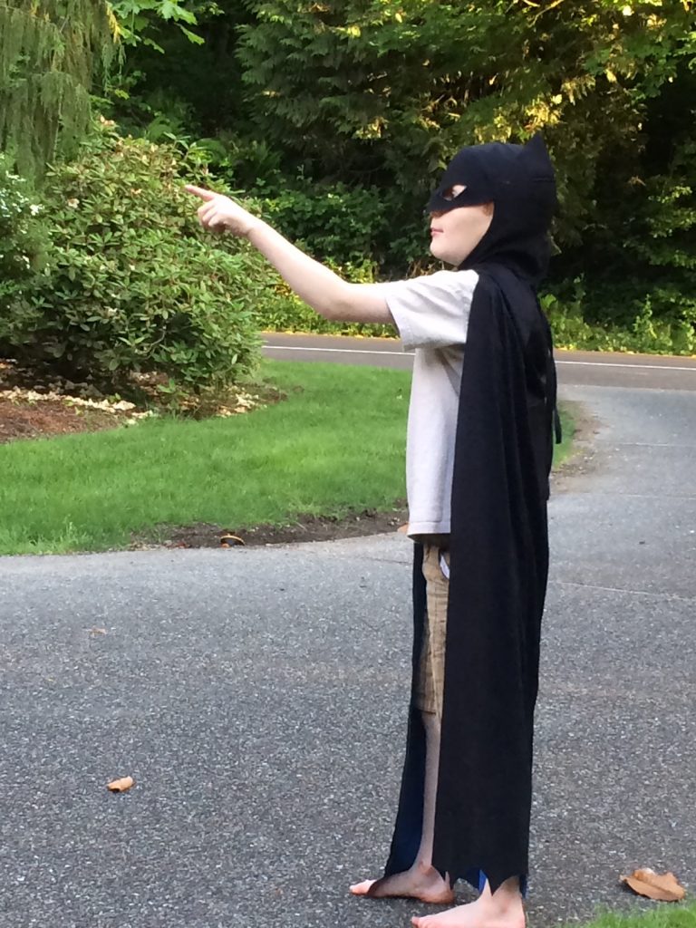 Nate dressed as Batman outside, trees in the background. Nate is standing sideways, pointing to something off the distance, out of the frame..