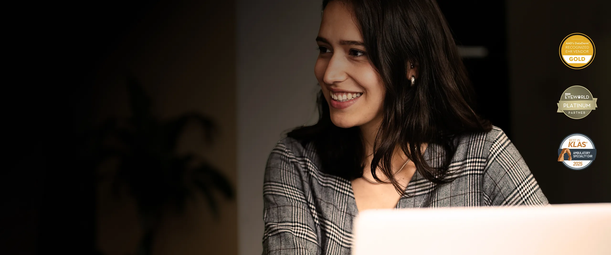 Smiling woman with dark hair in plaid blazer working on a laptop with award badges on the right.
