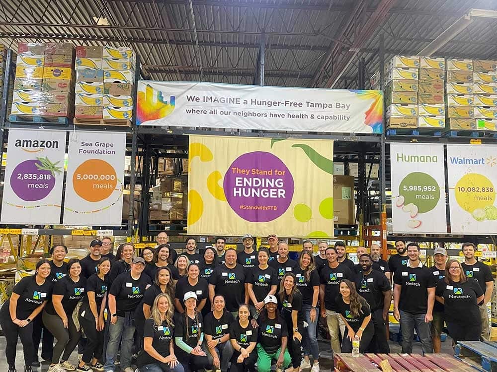 Group of people wearing Nextech shirts posing in a warehouse with banners about ending hunger and meal donations.