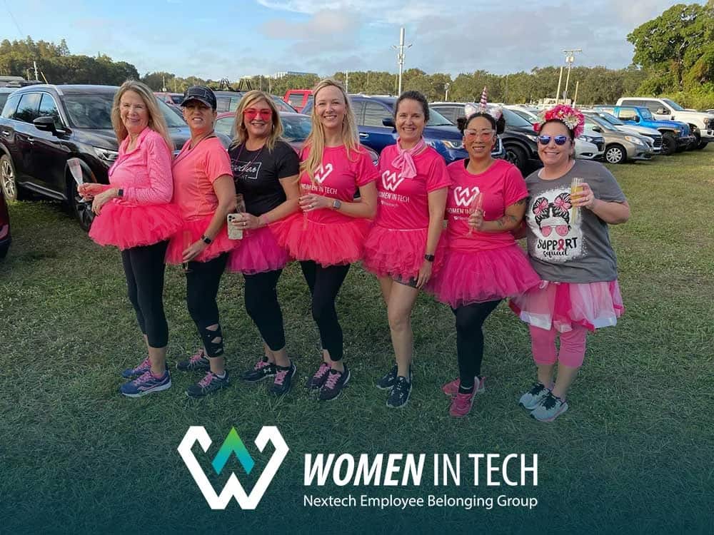 Seven women wearing pink tutus and shirts posing outdoors with cars behind them at a Women in Tech event.