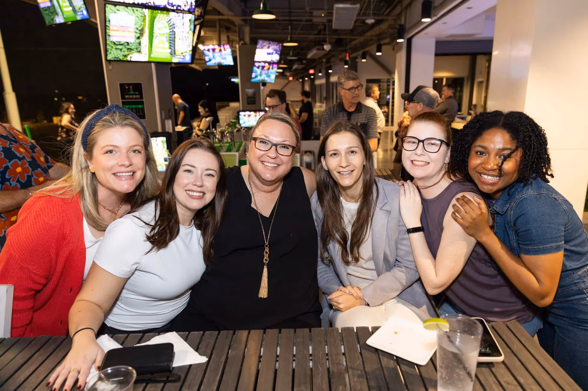 Six women smiling and posing closely together at a wooden table inside a lively venue.