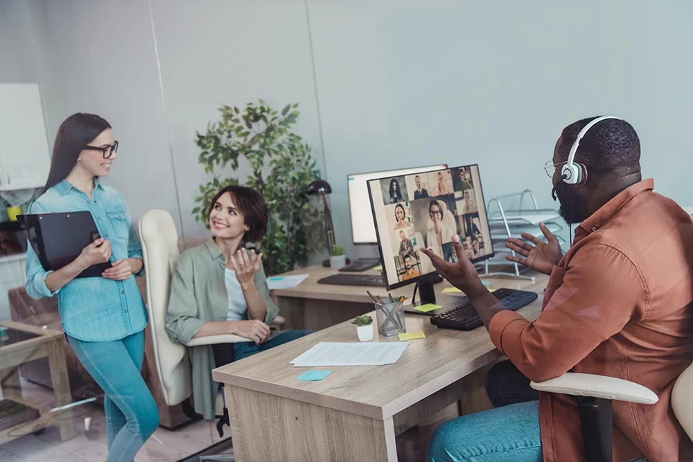 Three coworkers in an office, one on a video call with multiple people on screen, two others talking beside desk.