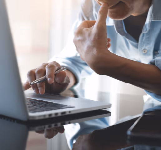 Person in blue shirt holding a pen, looking thoughtful while working on a laptop.