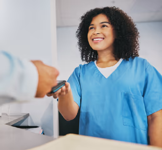 Smiling healthcare worker in blue scrubs handing a device to another person.
