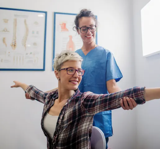 Physical therapist assists woman with outstretched arms stretching in a clinical room with anatomy posters.