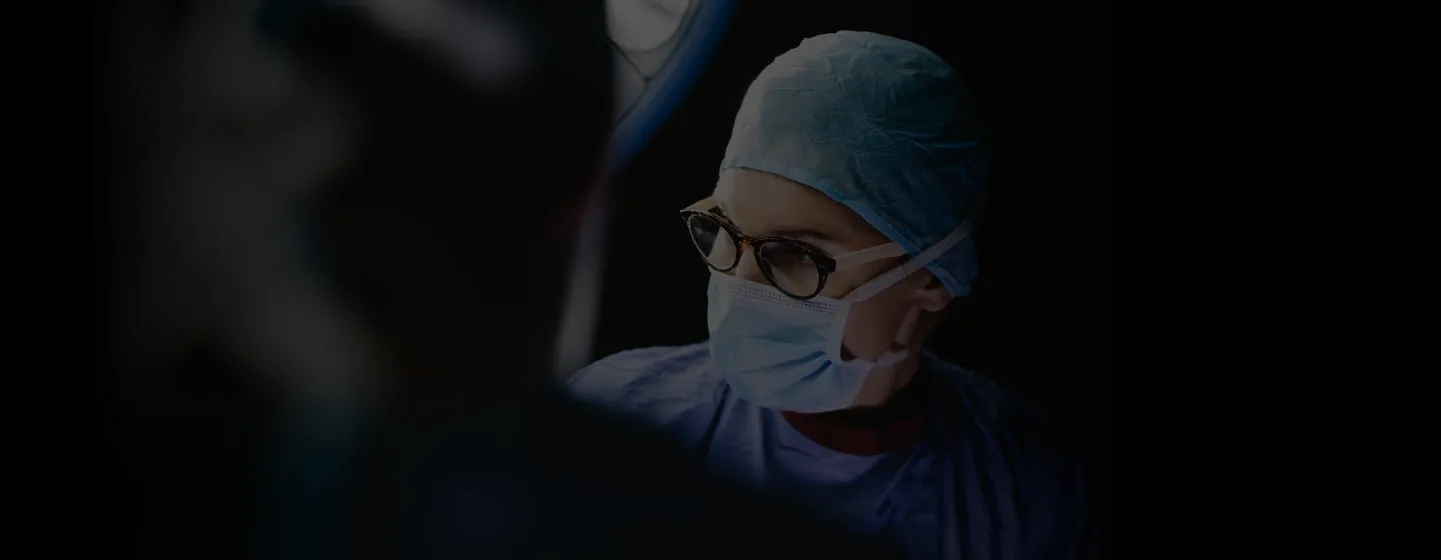 Medical professional wearing glasses, surgical mask, and cap in a dimly lit room.