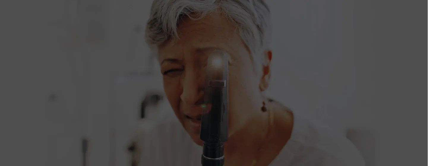 Older woman with short grey hair examining her eye using a handheld medical device with a light.