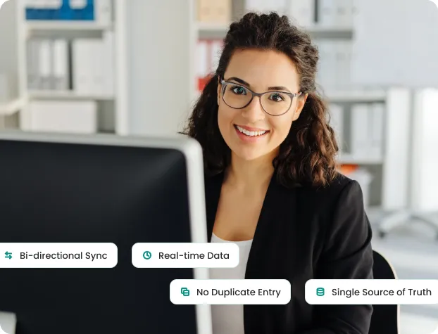 Smiling woman with glasses sitting at computer in office highlighting data sync and no duplicate entries.