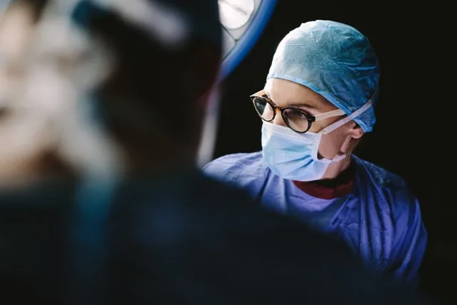 Surgeon wearing glasses, a surgical mask, and cap, looking to the side during an operation.