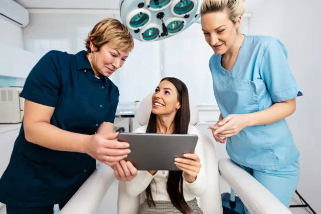 Two healthcare professionals showing a tablet to a smiling female patient seated in a medical office chair.