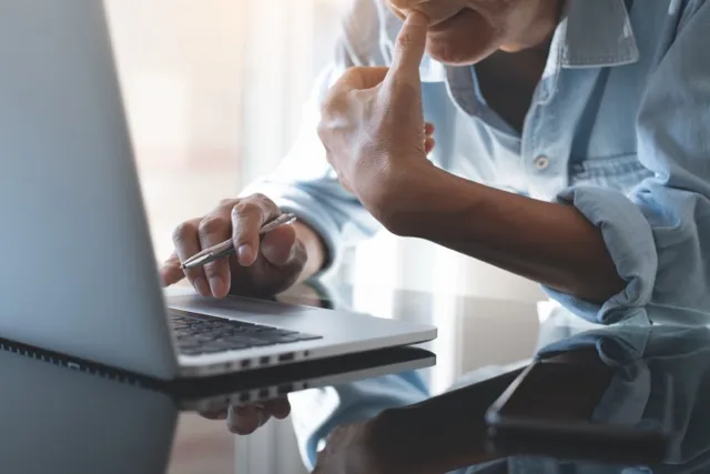 Person in denim shirt working on laptop with a pen in hand and smartphone on glass table.