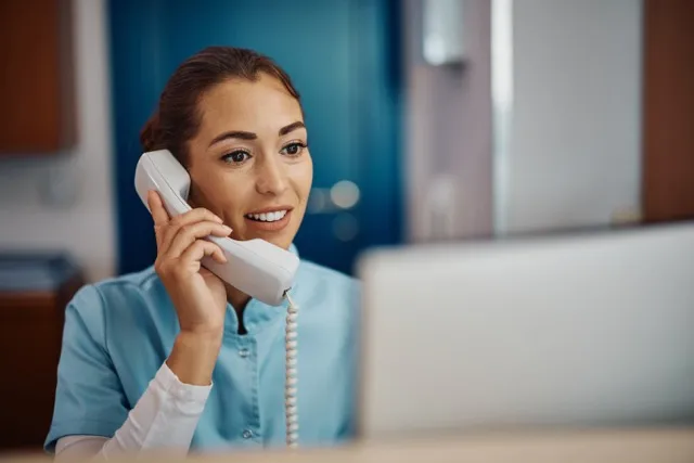 Smiling woman in blue scrubs talking on a corded phone at a reception desk with a computer.