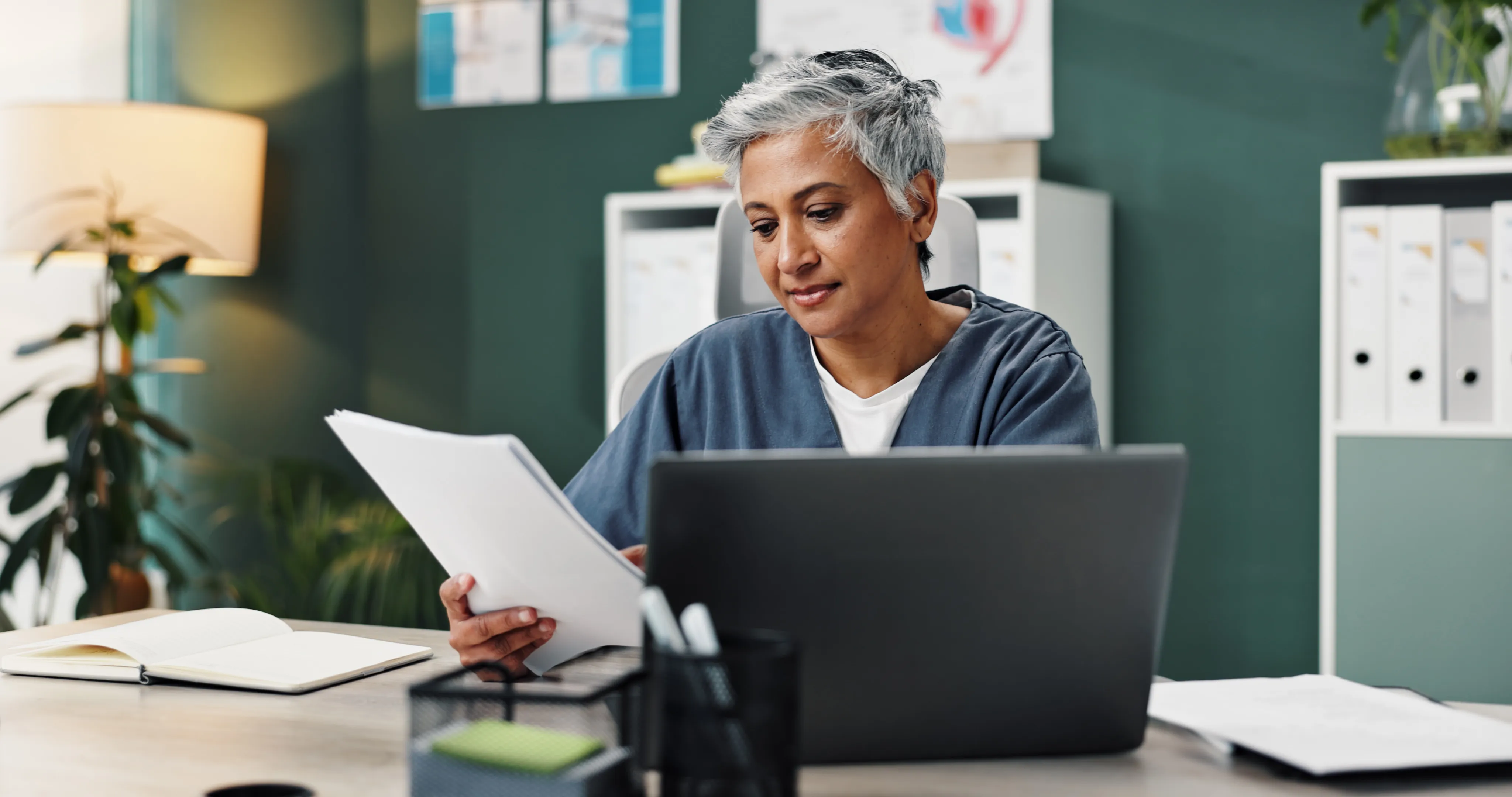 Woman with short gray hair reviewing papers at a desk with laptop and office supplies.