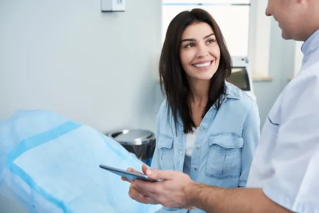 Smiling woman in denim shirt talking to a healthcare professional showing something on a tablet.