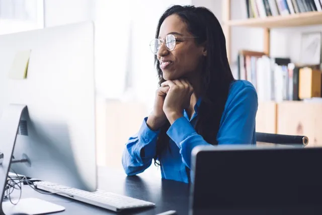 Woman wearing glasses and blue shirt smiling while looking at a computer screen in an office.