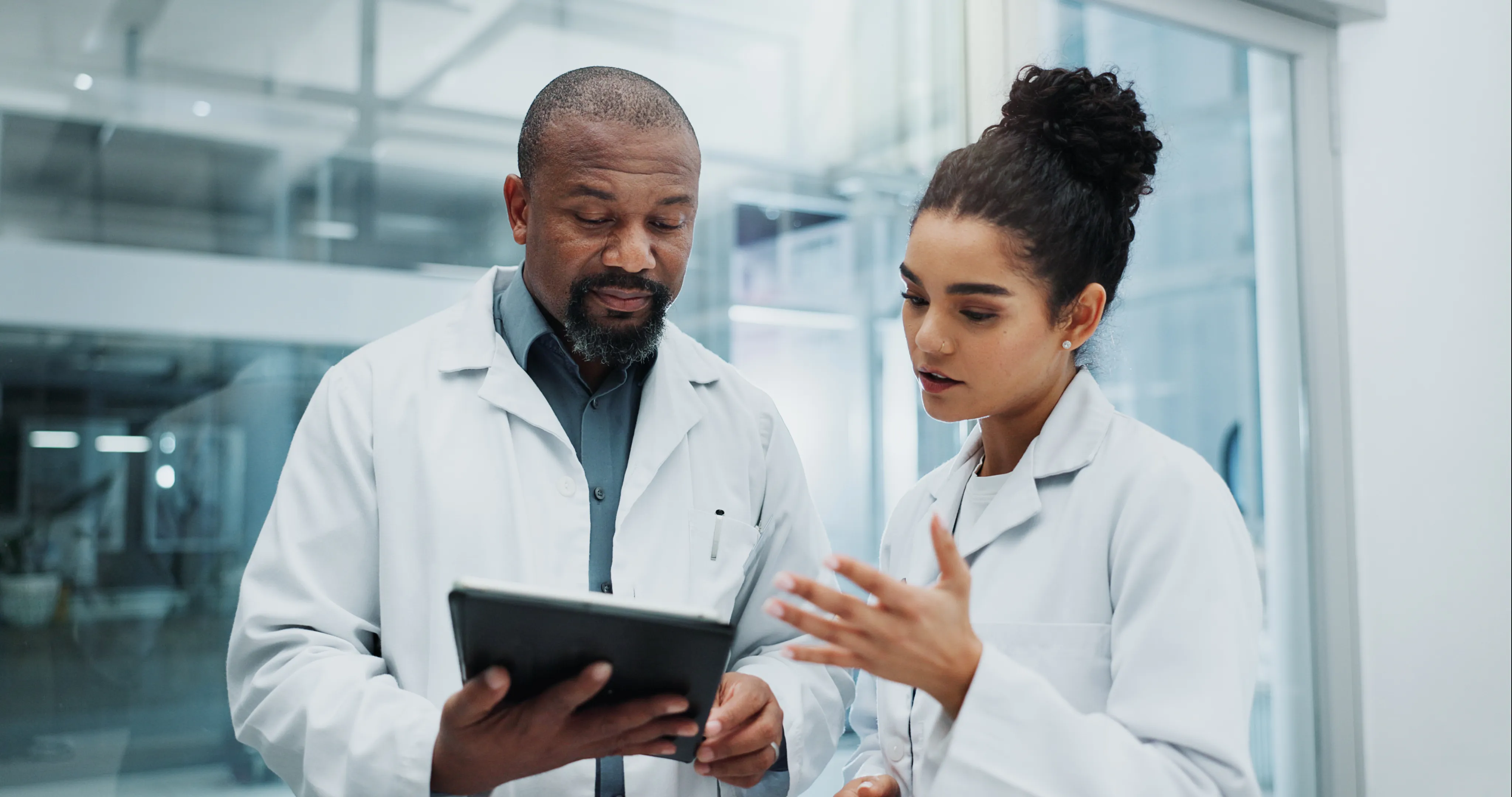 Two scientists in white lab coats discussing data on a tablet in a modern laboratory.