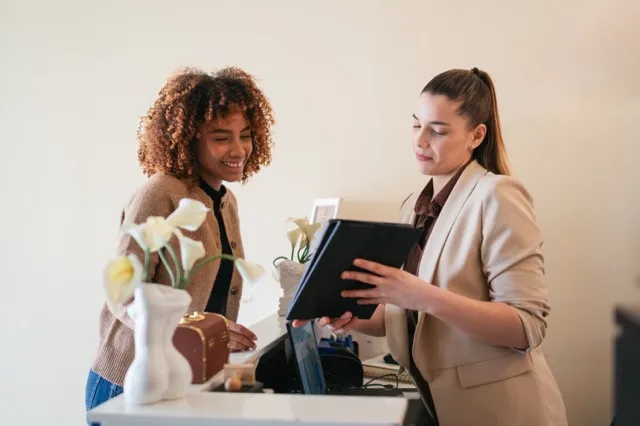 Two women at a counter, one showing a tablet to the other, with a vase of flowers on the counter.