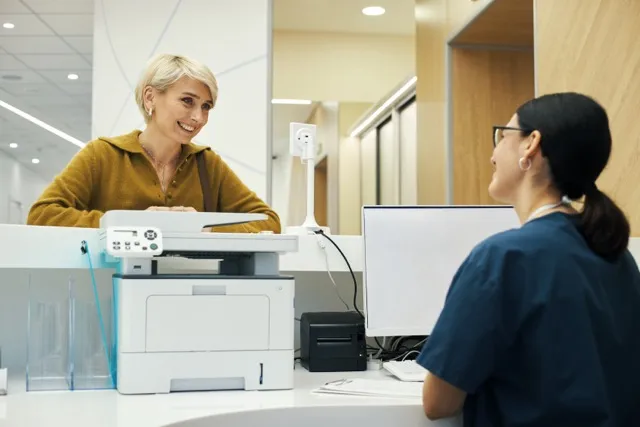 Smiling woman in mustard shirt talks to seated receptionist at a modern office desk with printer.