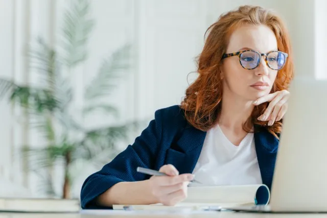 Red-haired woman wearing glasses writing in a notebook while looking at a laptop screen.