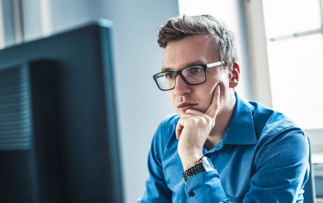 Young man wearing glasses and blue shirt focused on a computer screen in a bright room.