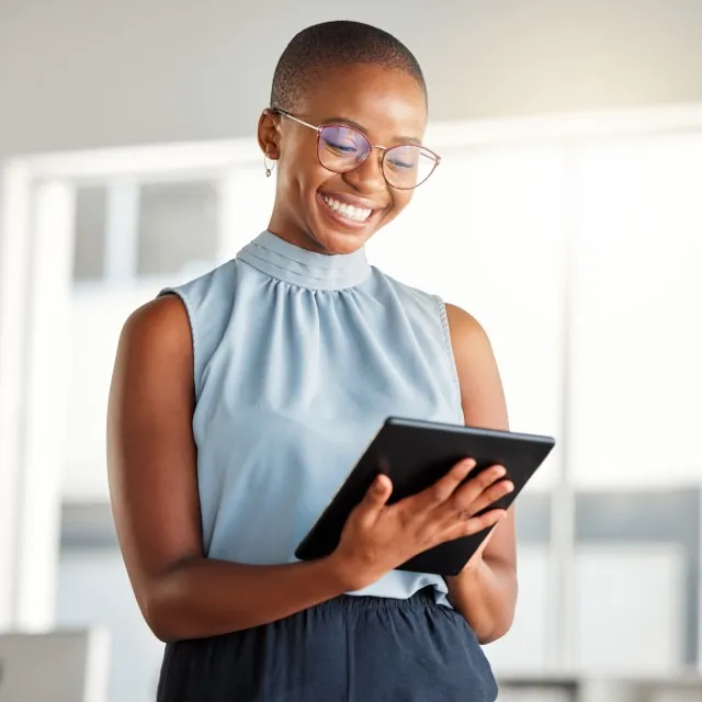 Smiling woman wearing glasses looks at a tablet in a bright office setting.