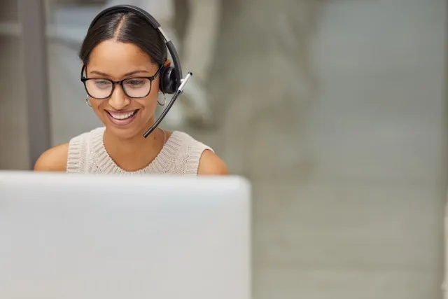 Smiling woman wearing headset and glasses, looking at a laptop in a bright indoor setting.