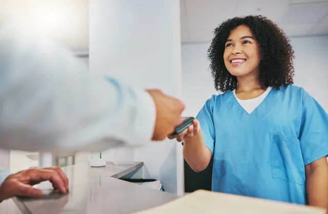 Smiling woman in blue medical scrubs handing a smartphone to a person across a counter.