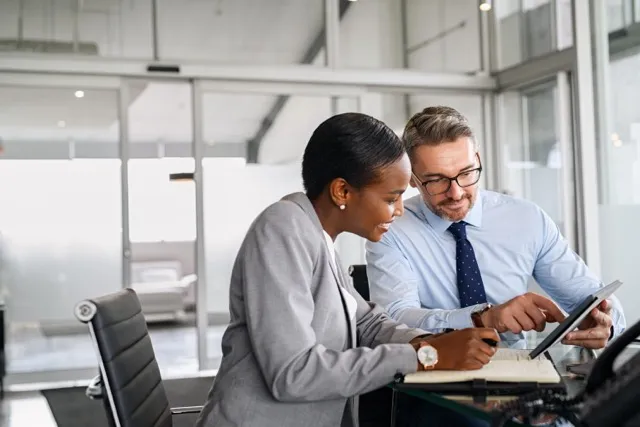 Two business colleagues review information on a tablet together in a glass-walled office.