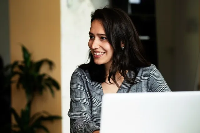 Smiling woman with long dark hair sitting behind a laptop in a softly lit room.