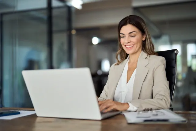 Smiling woman in beige blazer working on a laptop at a wooden desk in an office.