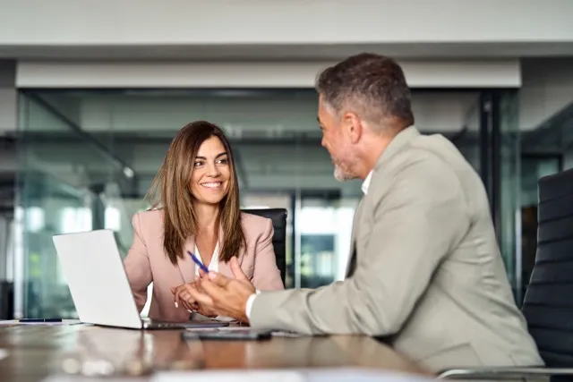 Smiling businesswoman talks with male colleague across a table with laptop in modern office.