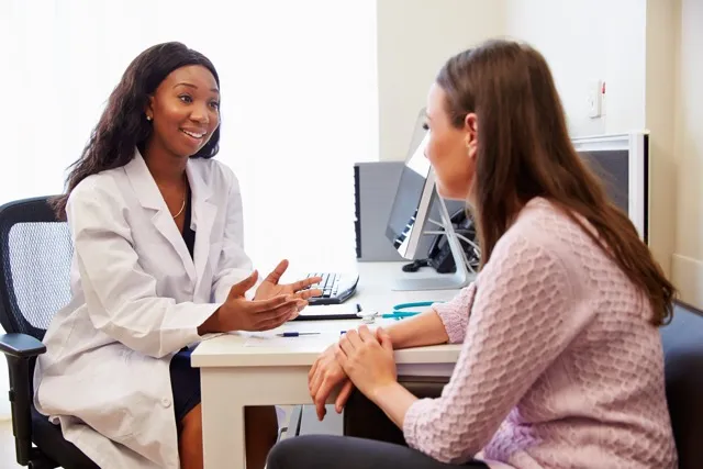 Female doctor in white coat talking to a female patient sitting across a desk in a medical office.