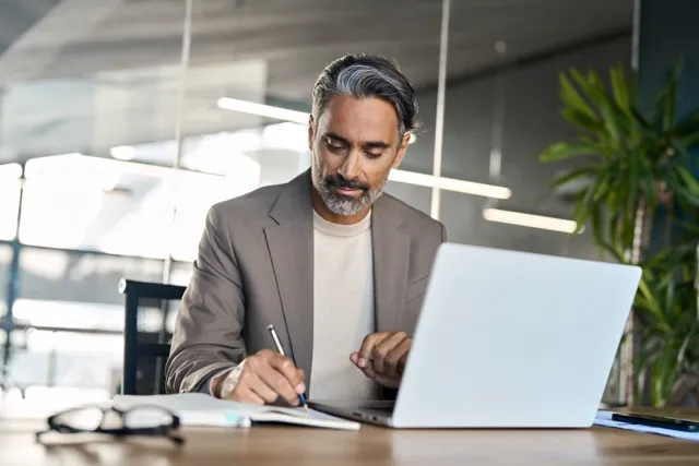 Man in blazer writing in notebook while sitting at desk with open laptop in bright office.