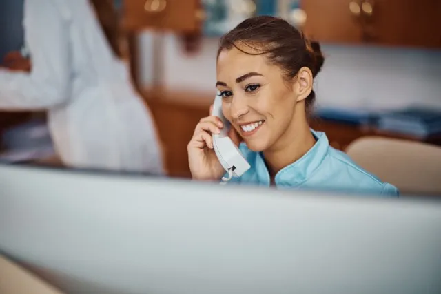 Smiling woman in a blue shirt talking on a white telephone at a desk in an office.