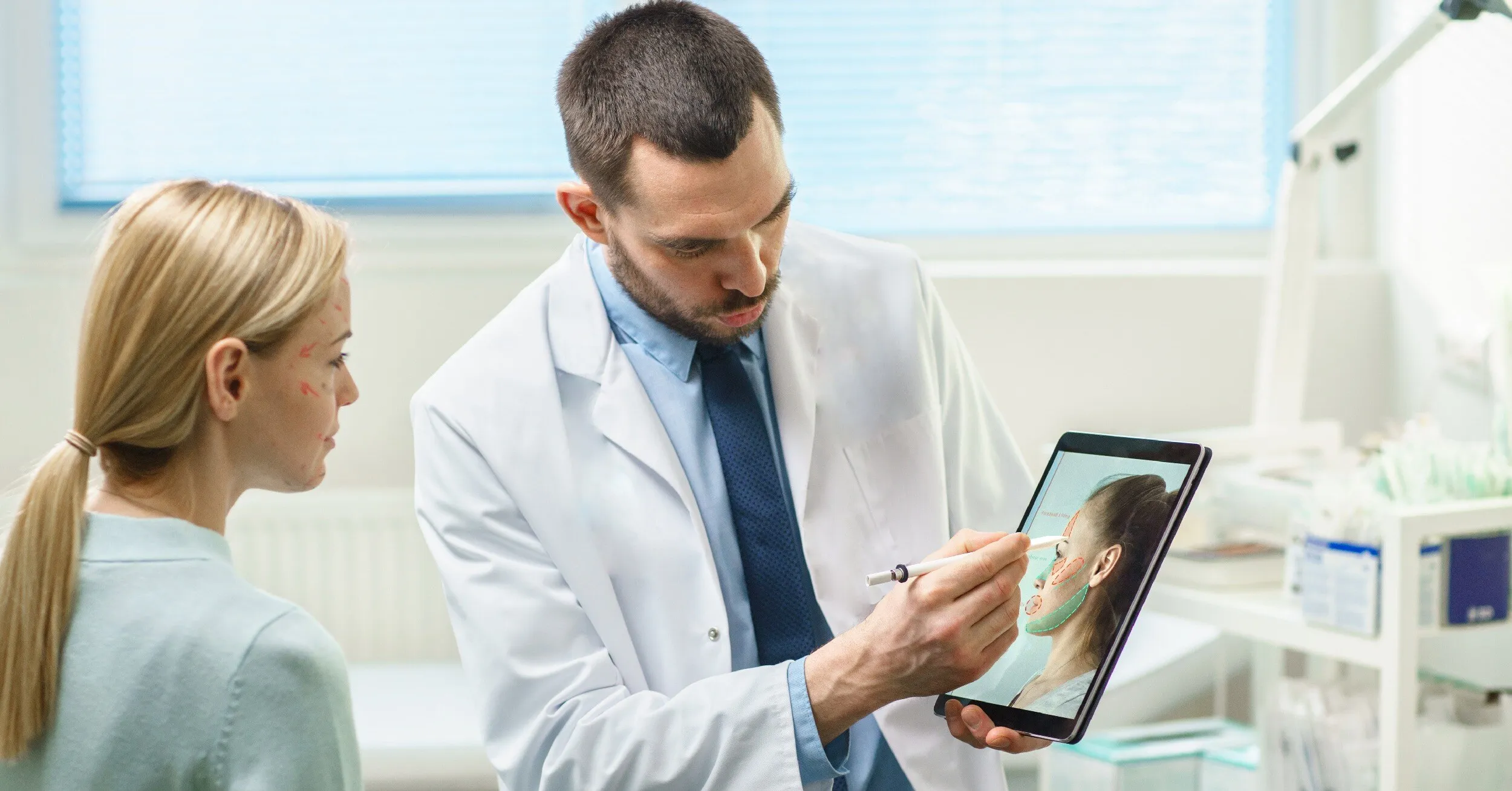 Doctor shows a woman marked facial areas on a tablet for plastic surgery consultation.